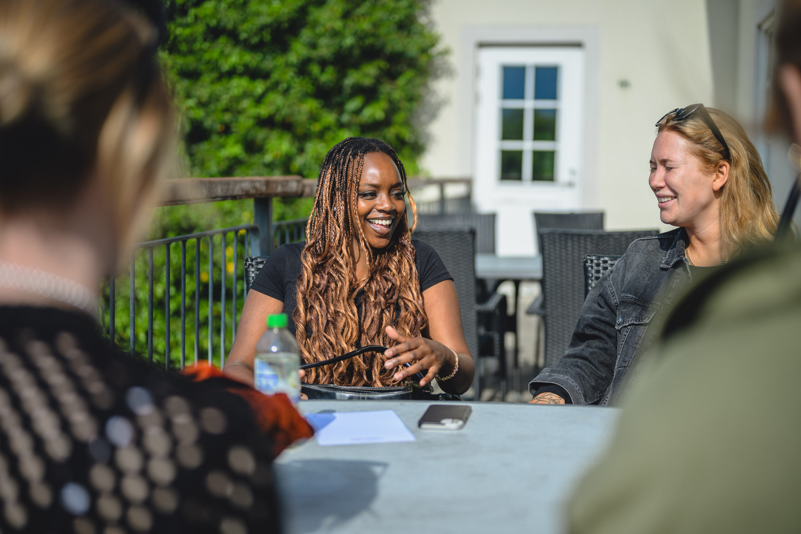 Deltagare ler och diskuterar något tillsammans med kurskamrater vid ett bord i solen utanför skolbyggnaden Herrgården